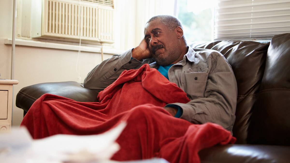 A man sitting on a couch under a blanket, leaning his head into his hand, looking dejected.