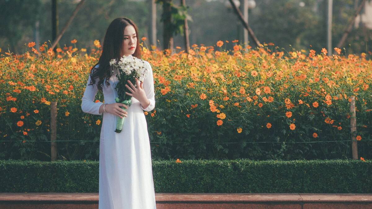 A woman in a white dress holding a bouquet of white flowers standing in front of a garden of orange flowers. She's looking to the side sadly and longingly.