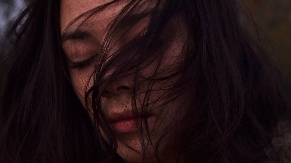 A close shot of a woman with her eyes closed, wind blowing hair across her face, looking sad.