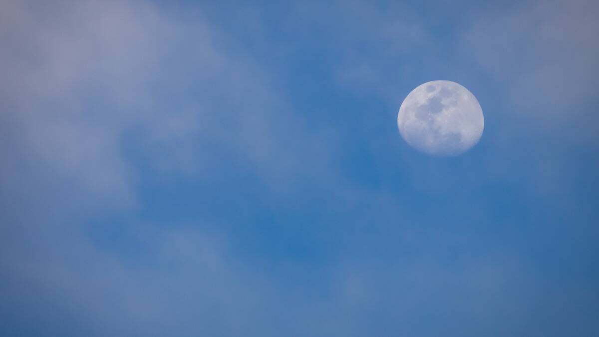 A nearly full moon in a blue sky, thin clouds seen surrounding it.