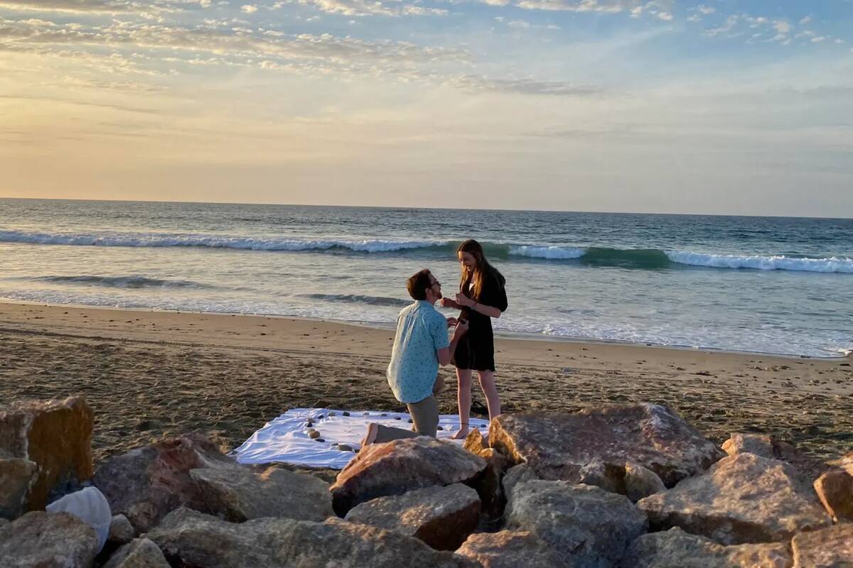 Tyler proposing to Kelsey on the beach, standing on a blanket with rocks arranged in a heart on it.