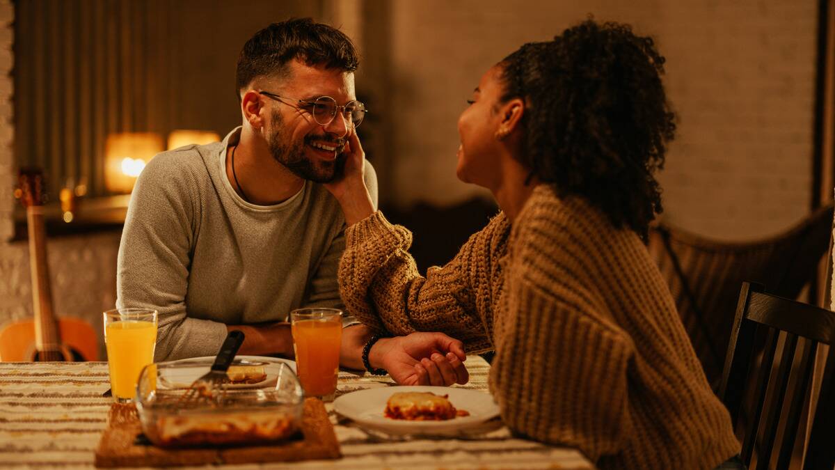 A couple sitting at a table for dinner, both smiling, the woman reaching out to touch the man's cheek.