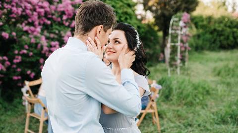 A couple standing in a garden, holding each other close, hands on each other's cheeks/jaw.