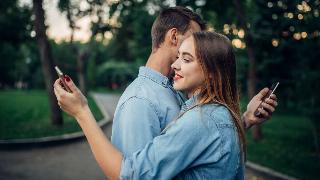 A photo hugging, chest to chest, but are each looking over the other's shoulder at their phone.