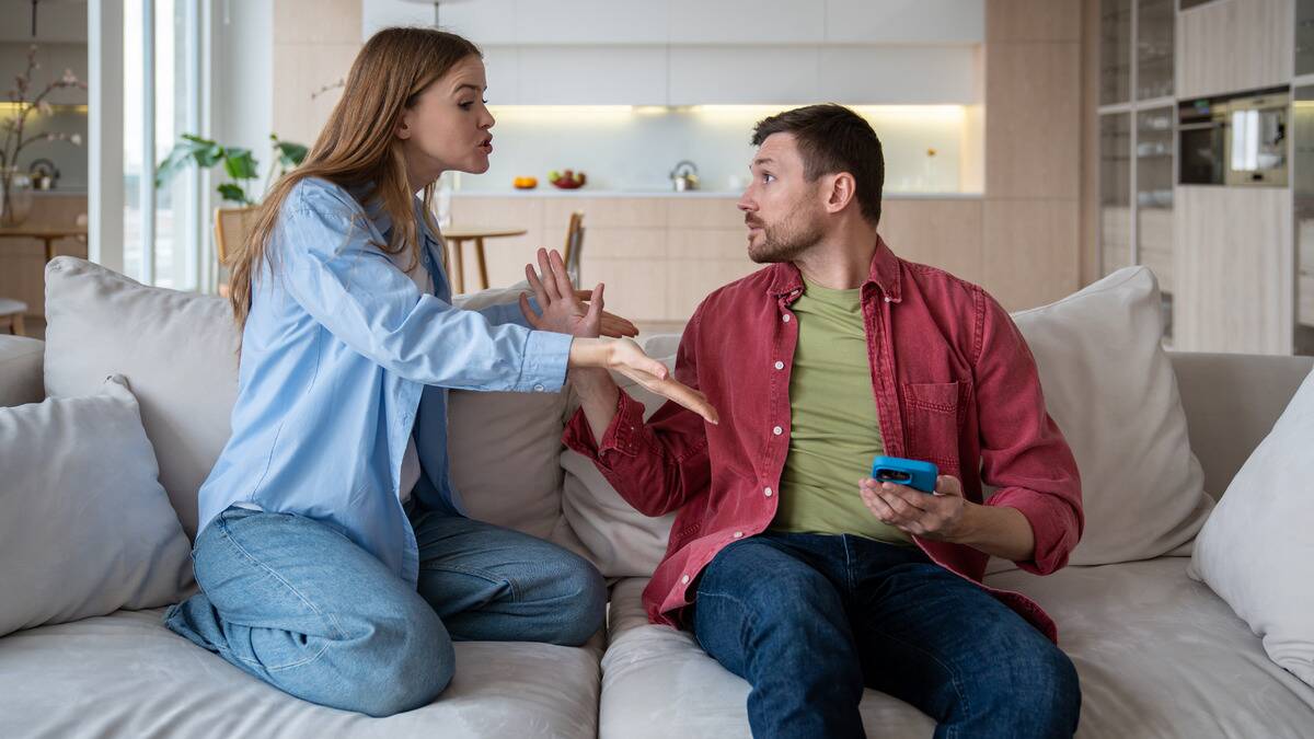 A couple arguing on the couch, the woman accusatorially pointing at the man's phone.