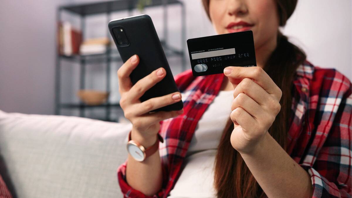 A close shot of a woman holding a phone in one hand, her credit card in another, putting in the numbers to make a purchase.