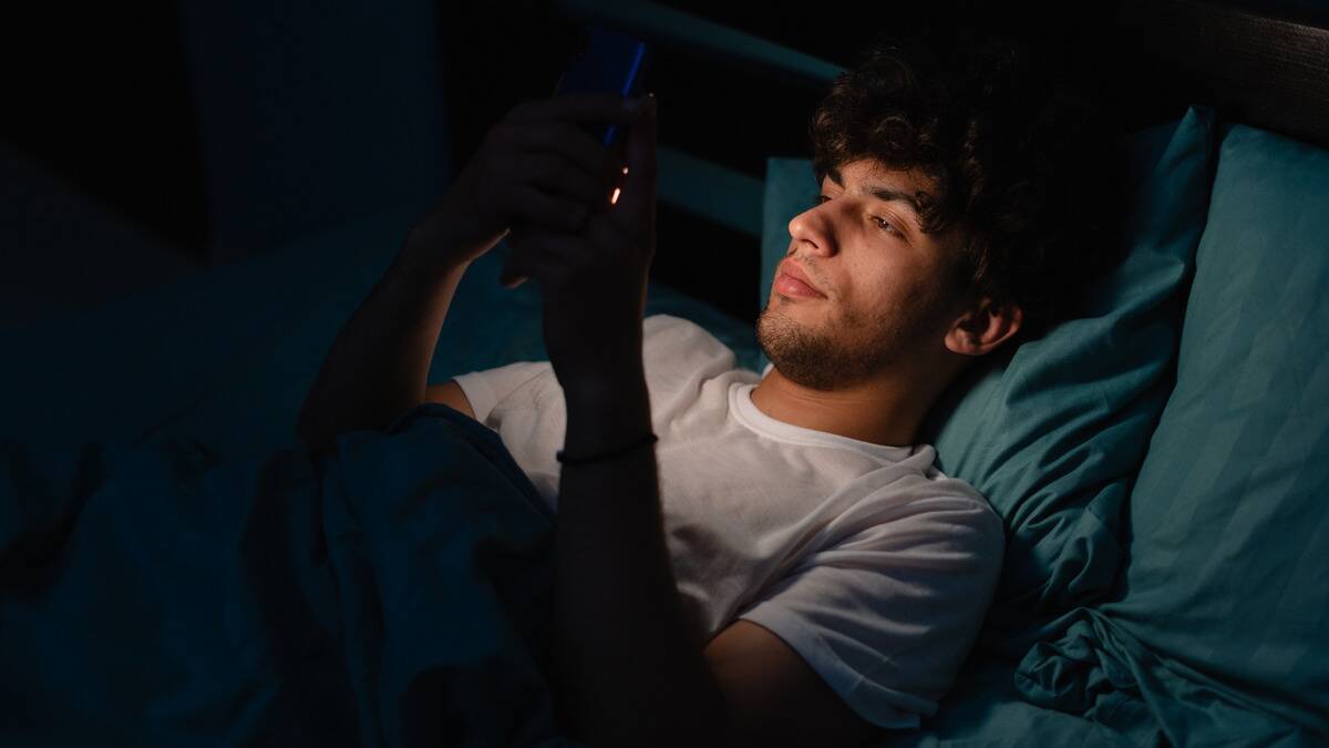 A man laying in bed in the dark, his face illuminated by his phone.