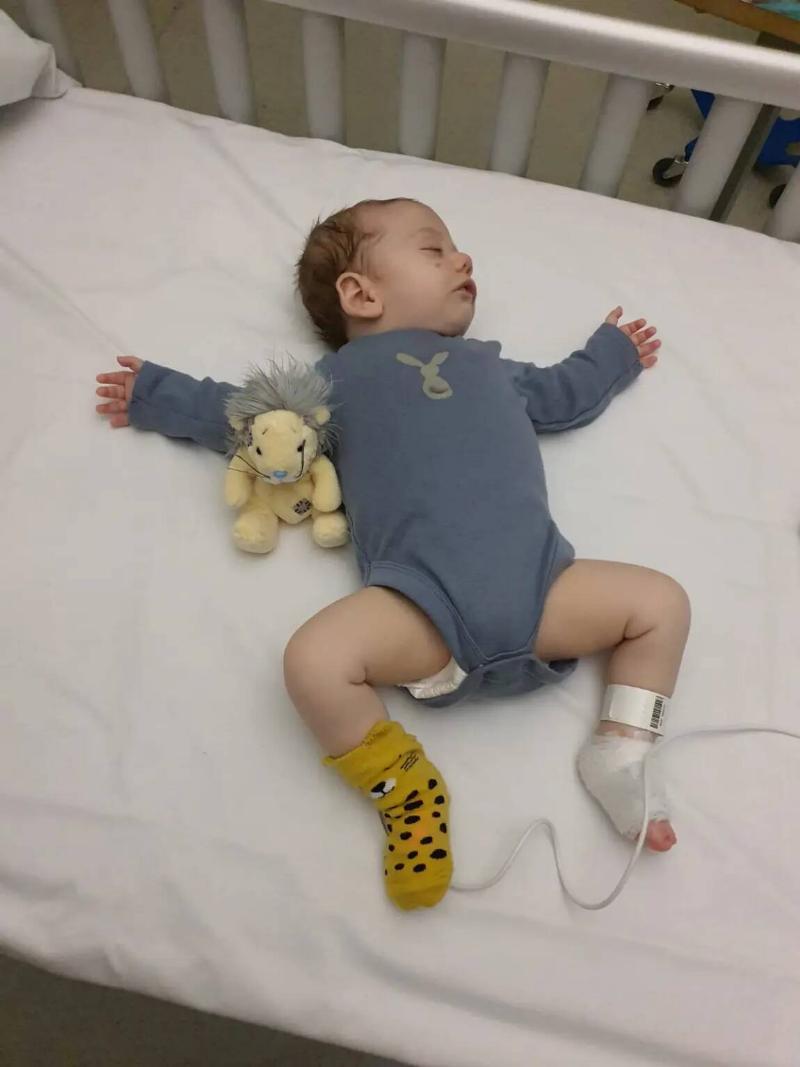 Baby Thomas in a hospital bed, a wire attached to his leg, splayed out in a blue onesie as he sleeps.