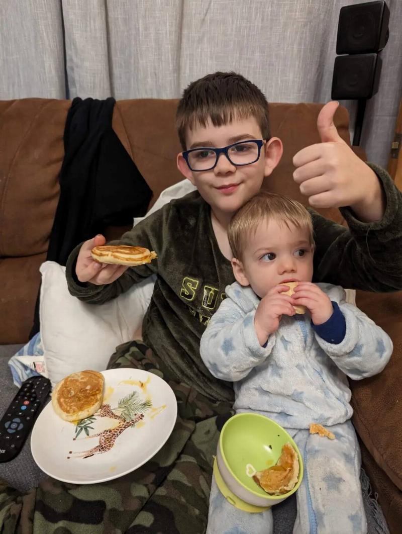 Thomas sitting in his older brother Lucas' lap as they both eat pancakes, Lucas giving a thumbs up.