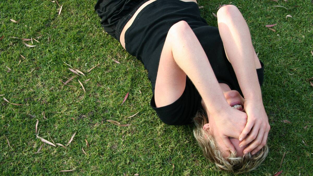 An upside-down shot of a woman laying in the grass, covering her face with her hands, looking pained.