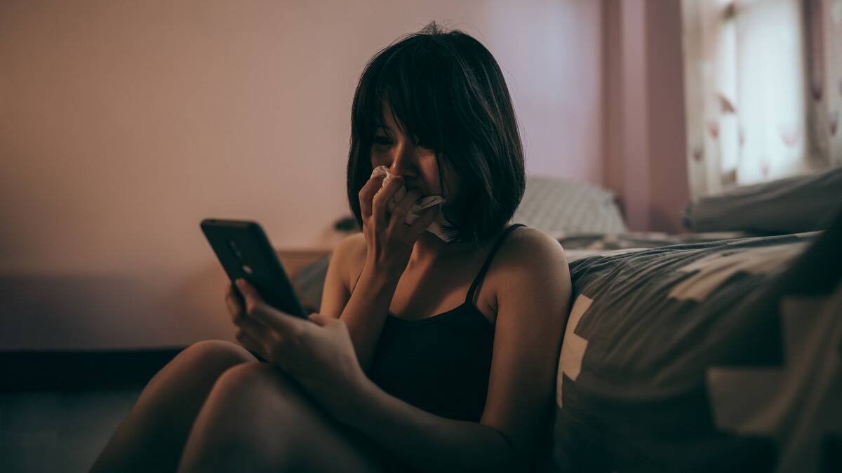 A woman sitting on the floor, leaning against her bed, crying as she looks at something on her phone, holding a tissue to her face.
