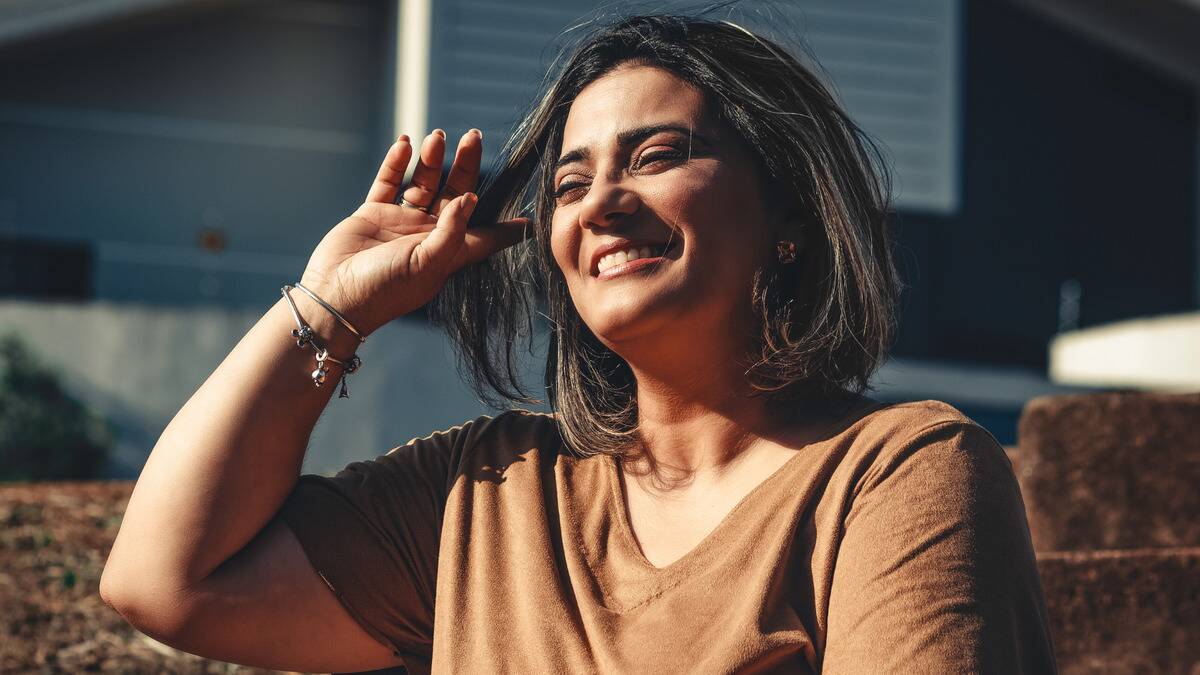 A woman smiling as she stands in the sunshine, brushing her hair away with her hand.