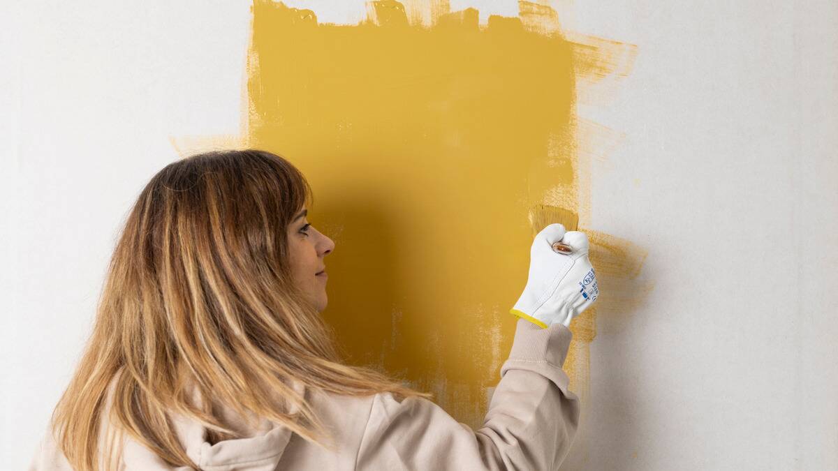 A woman painting her wall yellow with a small brush.