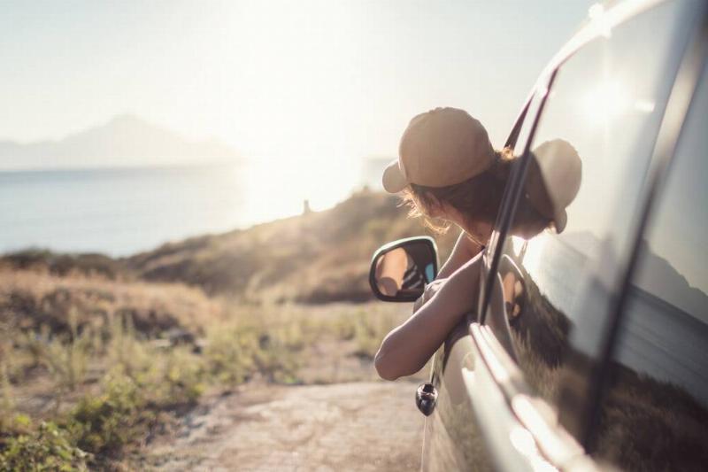 A woman leaning out the driver's side window of a car, looking out at the watery horizon she parked in front of.