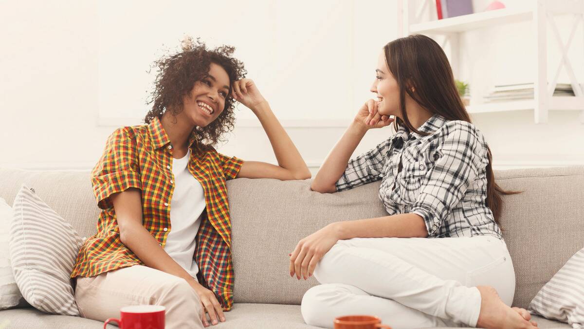 Two women smiling as they sit on a couch next to each other and chat.
