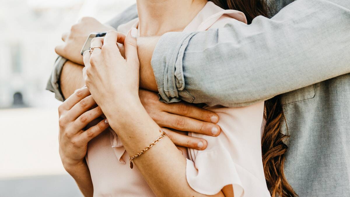 A close shot of a man hugging a woman from behind, her hands coming up to rest on his arms.