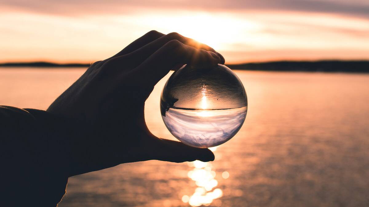 A hand holding a glass sphere in front of a body of water with the sun rising over it, that image reflected within the sphere.