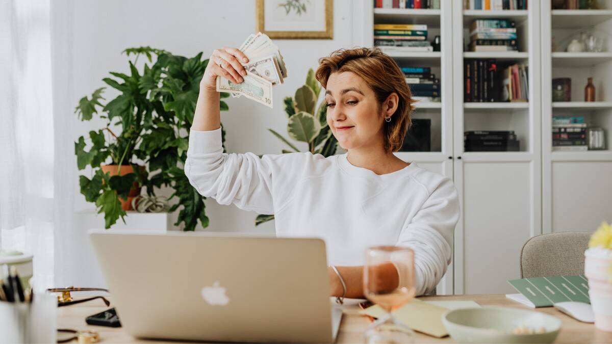 A woman sitting at her laptop, smiling as she holds up a fan of money in one hand.