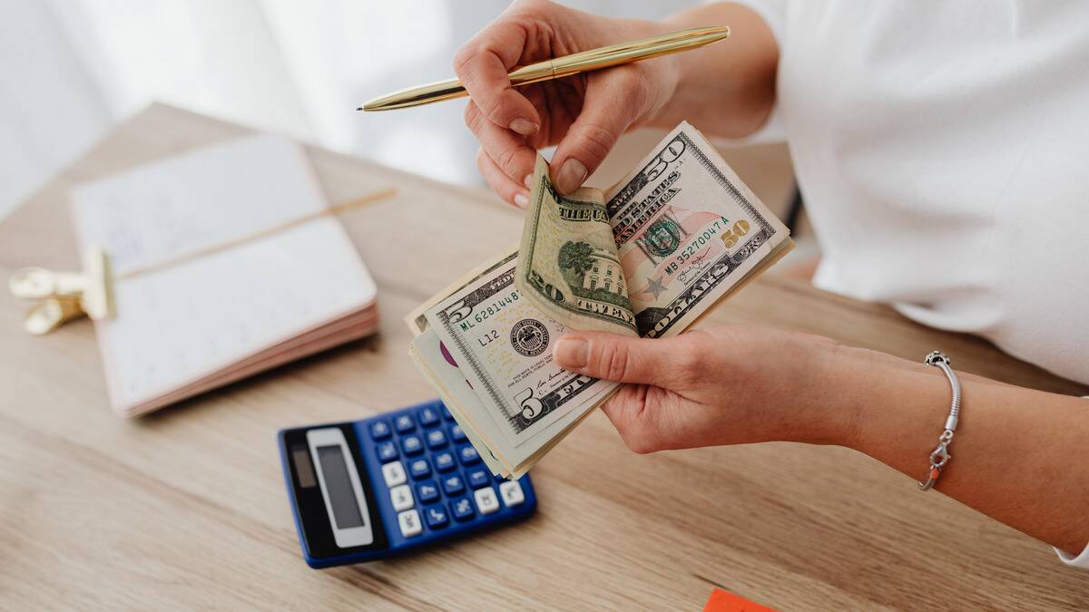 A pair of hands counting a stack of American bills, holding a pen with a notebook and calculator on the table below.