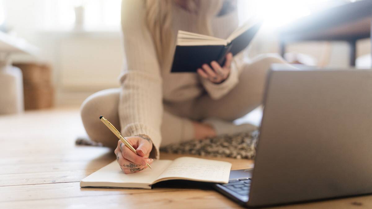 A woman sitting on the in front of a laptop, holding a book open in one hand and writing in a notebook with the other.