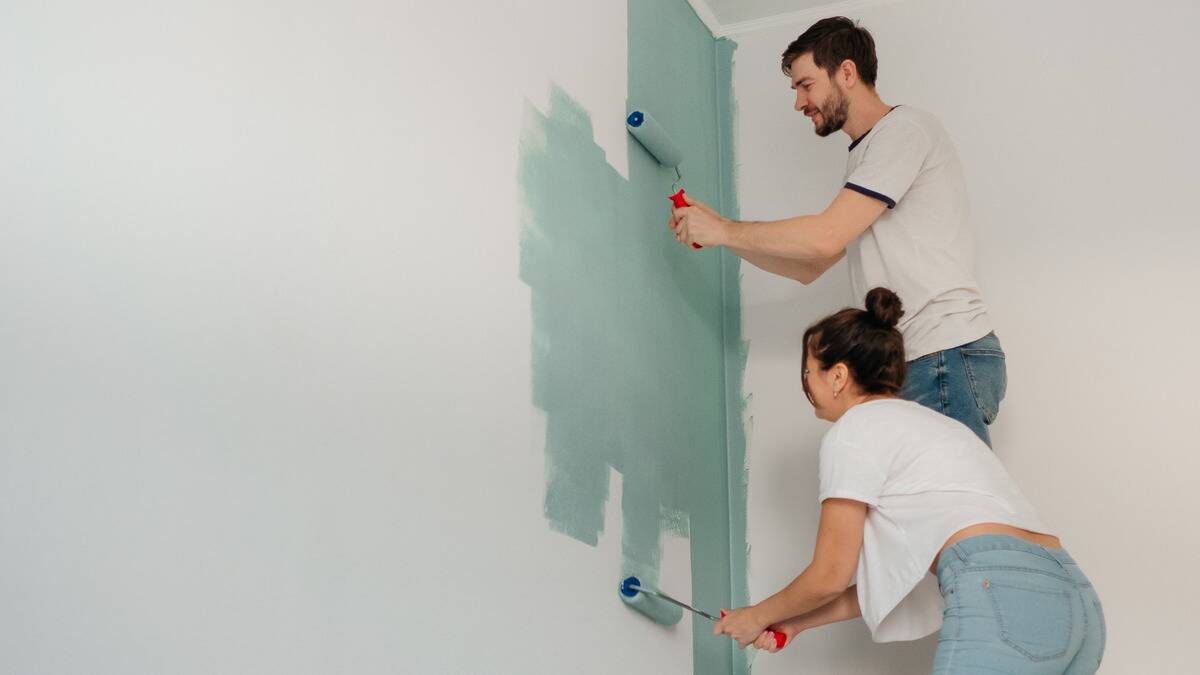 Two people, one on a step ladder painting a wall together from white to a light green.