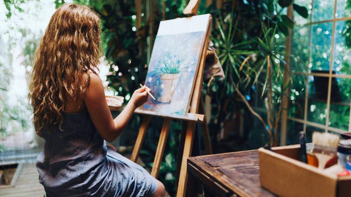 A woman sitting by greenery as she paints, back to the camera.