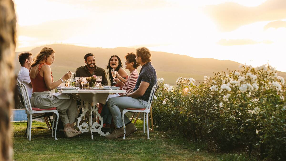 A group of friends sitting around a table outside, next to a garden of flowers, sun setting behind them.