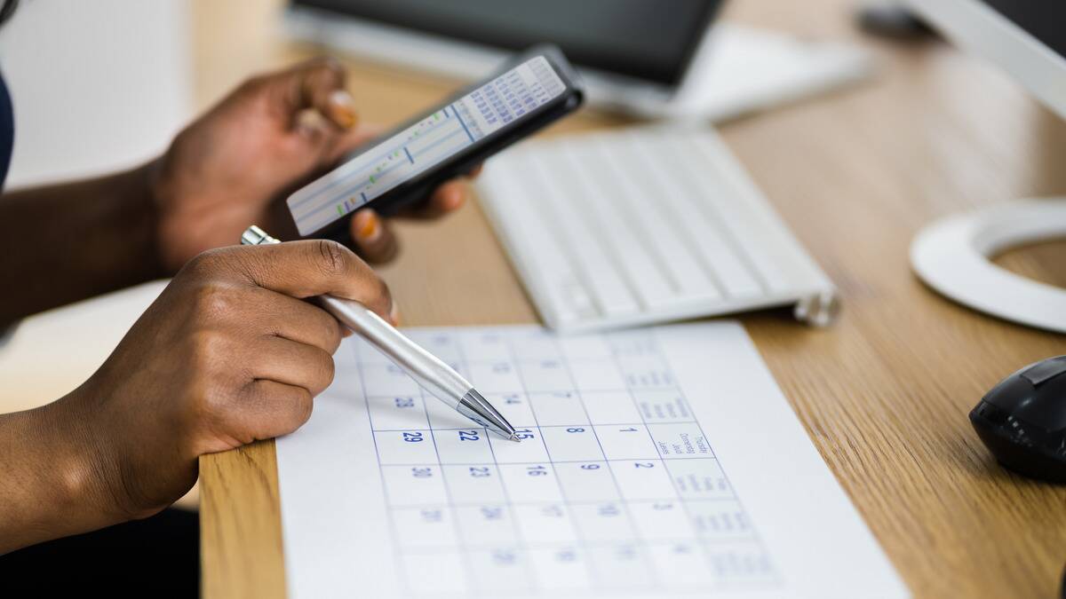 Someone holding a phone in one hand and writing on a large printed calendar in the other.