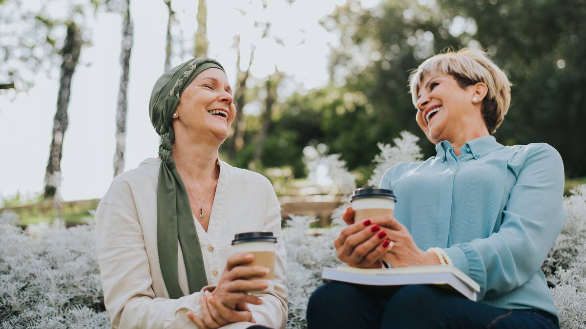 Two women sitting outside, laughing as they talk, both holding a cup of coffee.