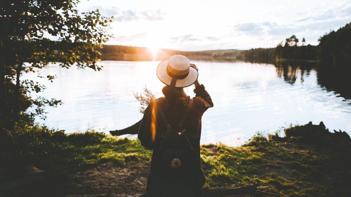 A woman standing by a lake at sunrise, a hand on the brim of her hat.