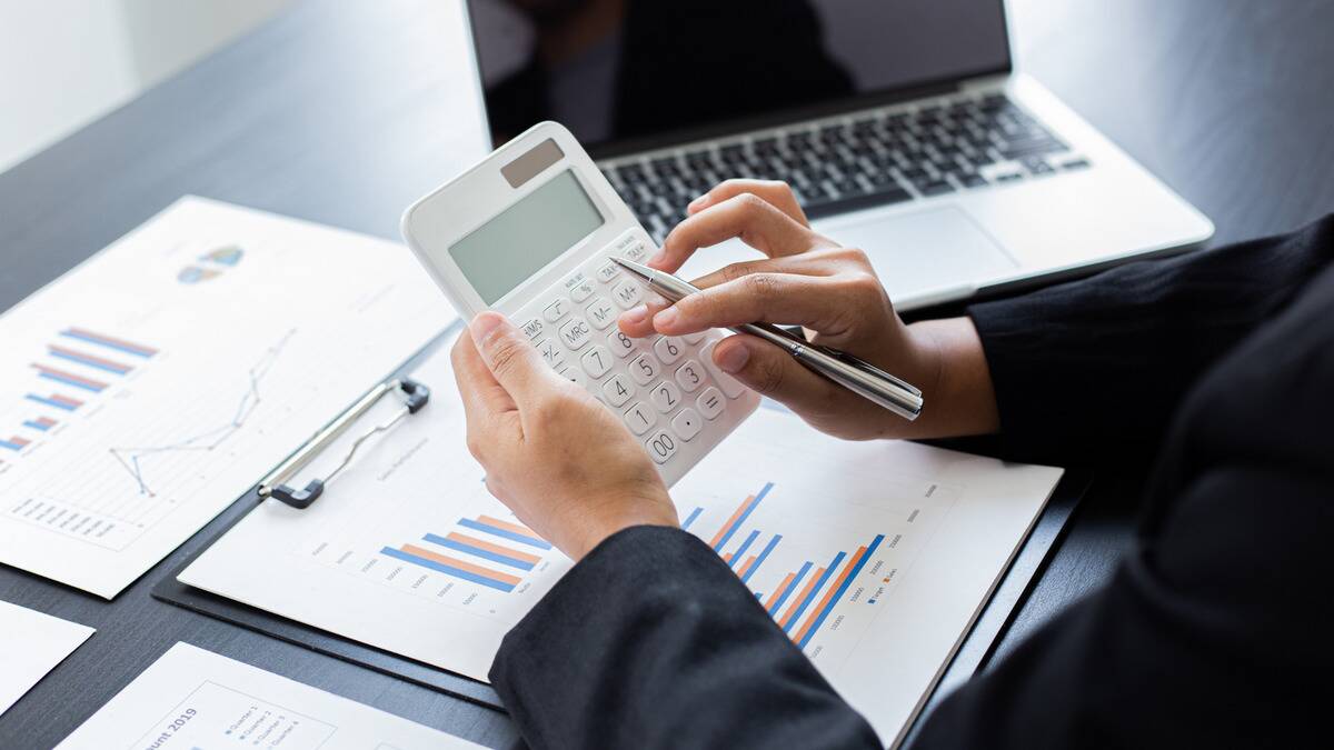 A pair of hands holding a calculator, punching in numbers, papers with graphs seen on the table below.