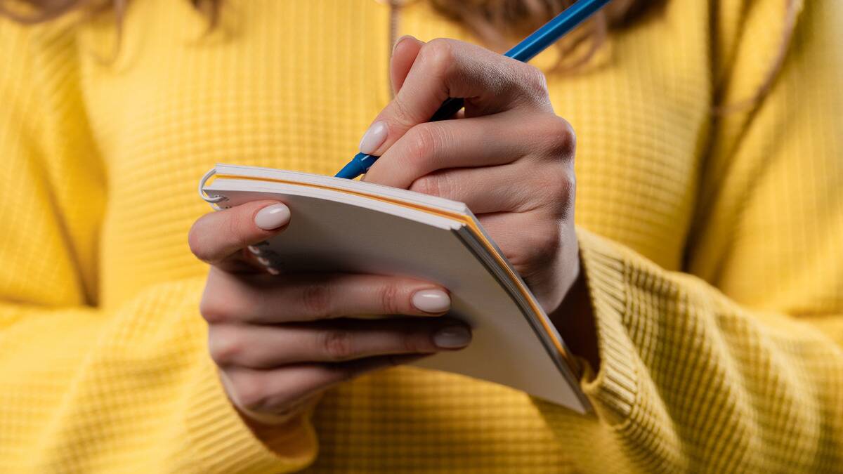 A close shot of a woman in a yellow sweater writing in a small notepad that she's holding.