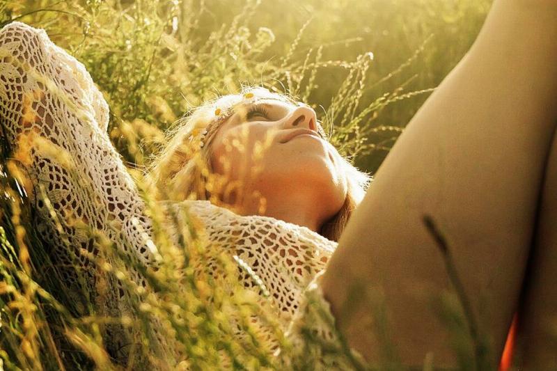 A close, low shot of a woman laying in the grass, hands behind her head, looking to the sky as the sun shines on her.