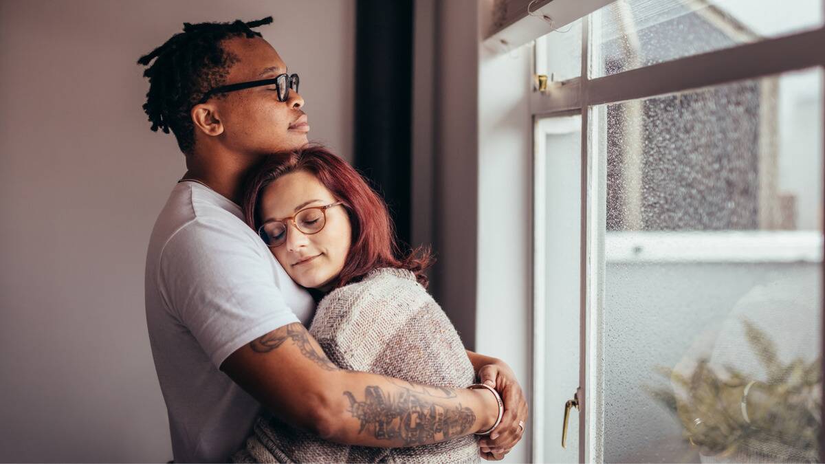 A couple standing in front of a window, hugging, both looking peaceful with their eyes closed.