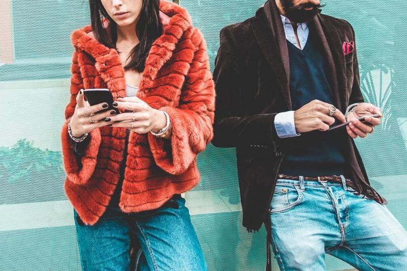 A photo of two young, trendy people, faces cut off in frame, showing off their outfits, both looking down at their phone in hand.