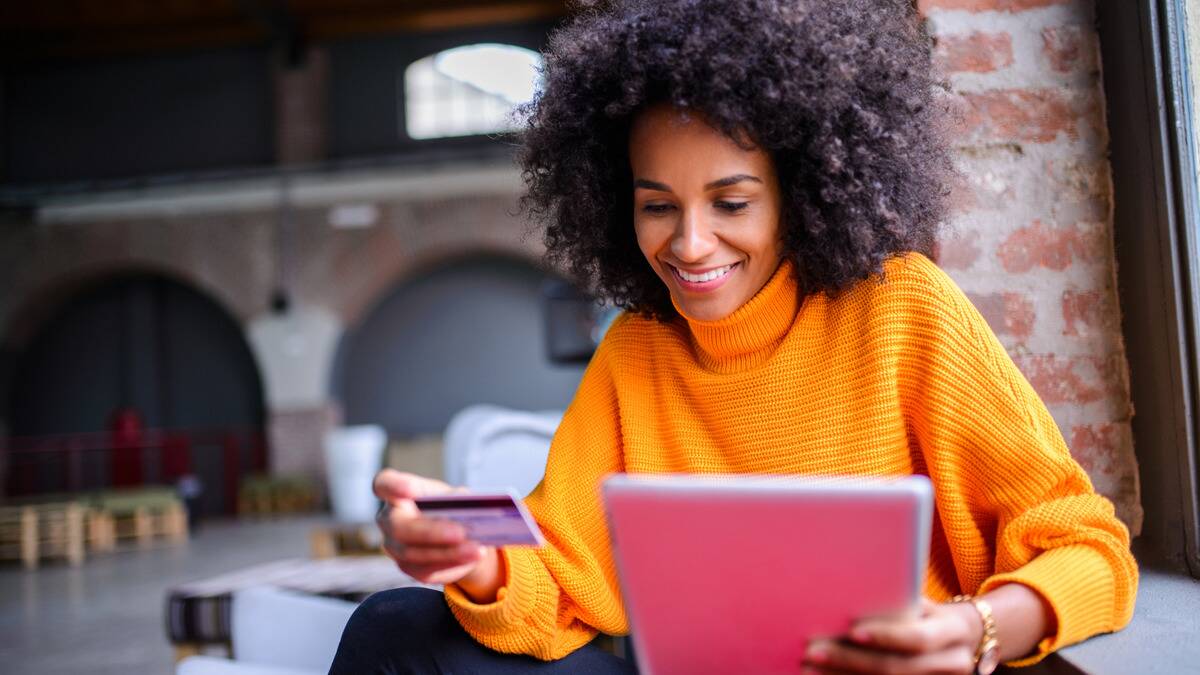 A woman holding her credit card in one hand, her iPad in the other, smiling as she buys something online.