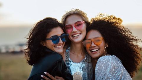 Three friends in colorful sunglasses posing close together, all hugging one another.