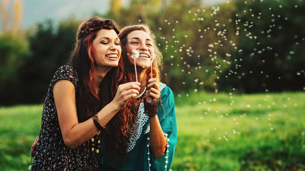 Two friends standing close side by side, each blowing dandelion seeds.