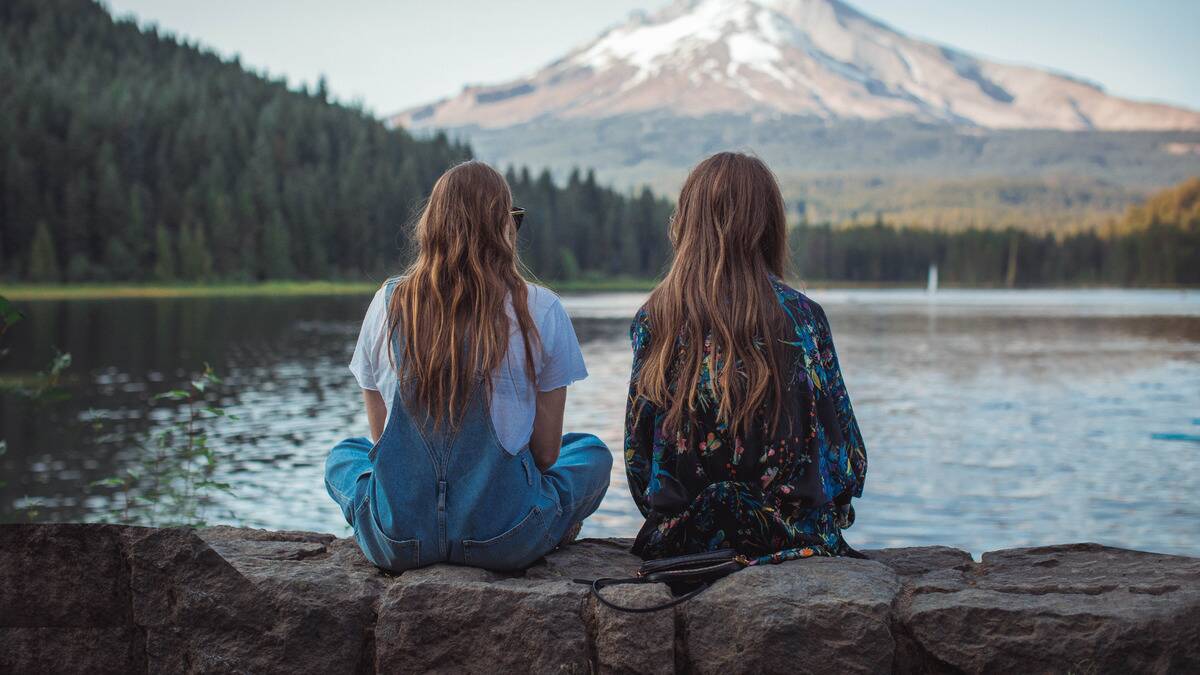 Two friends sitting outside in front of the water, facing away from the camera. 