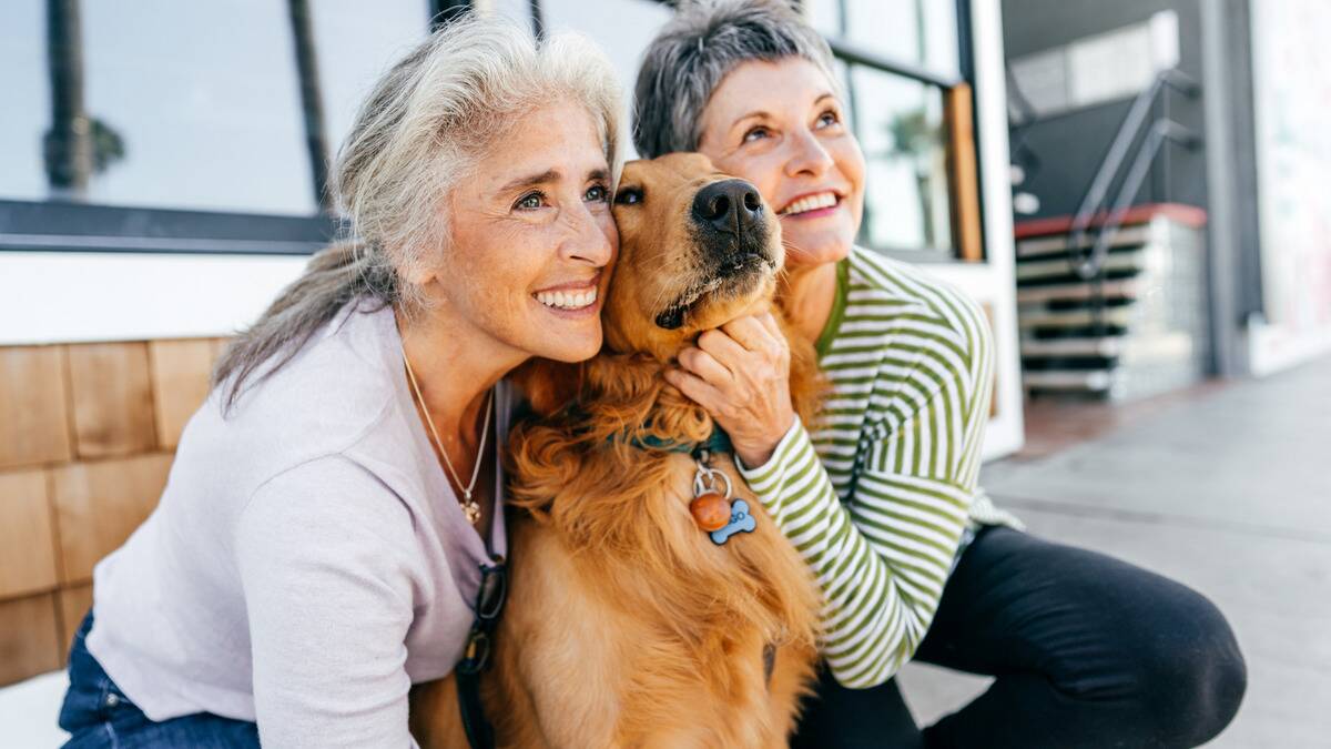 Two women outside on the either side of a golden retriever dog, both petting it and smiling.