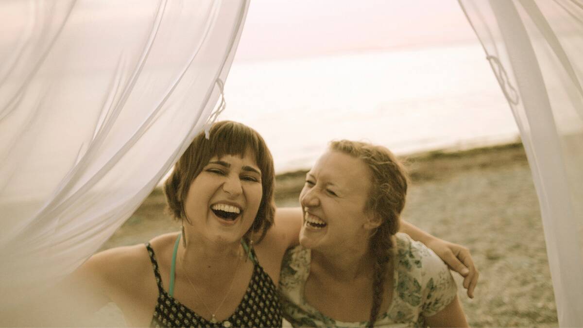 Two friends sitting next to each other and laughing on a beach, framed by billowing white fabric.