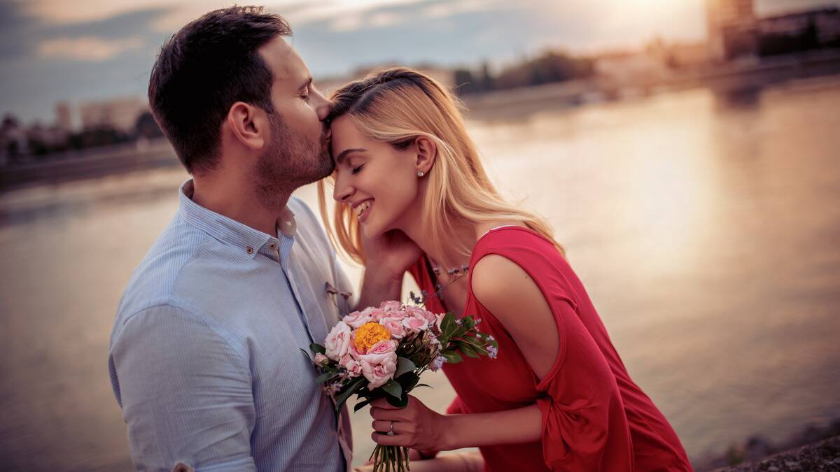 A tilted shot of a couple standing by a body of water, the woman holding a bouquet of pink flowers, the man kissing her forehead, both smiling with their eyes closed.