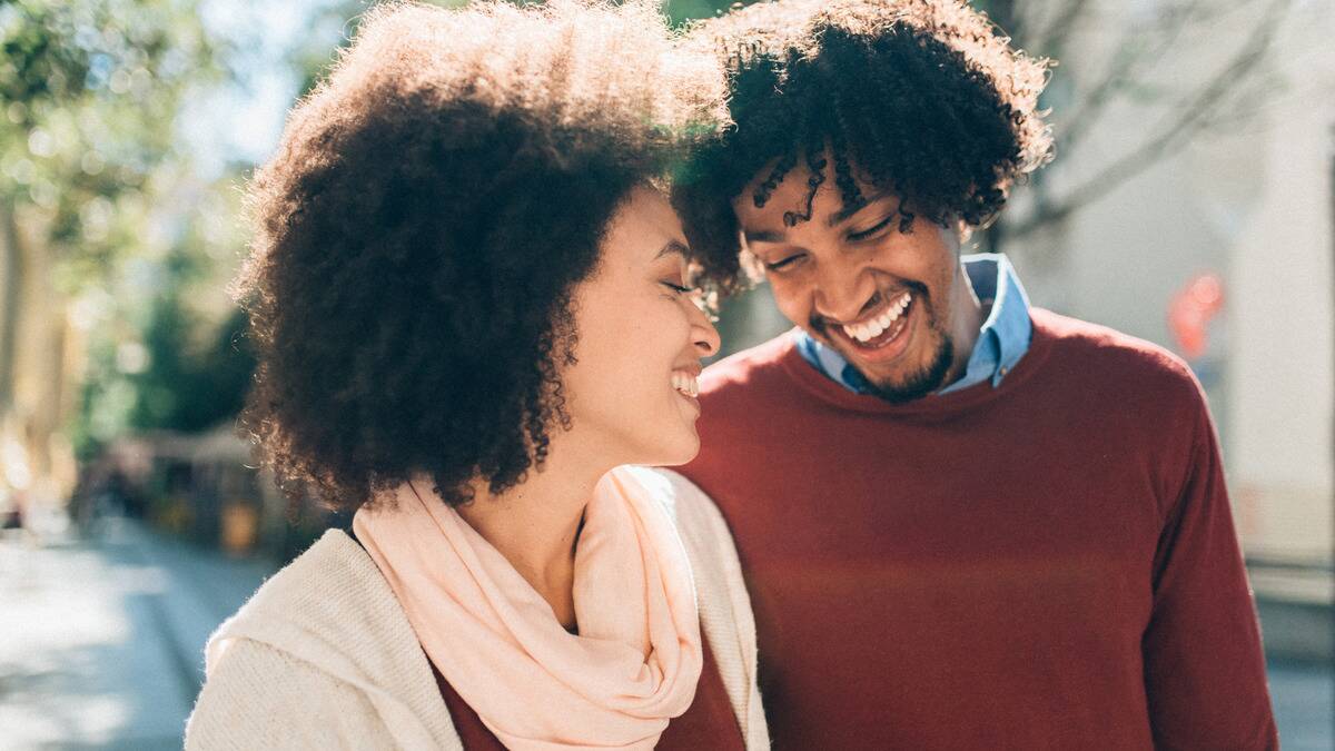 A couple walking down the street close, both laughing with their eyes closed as they talk.
