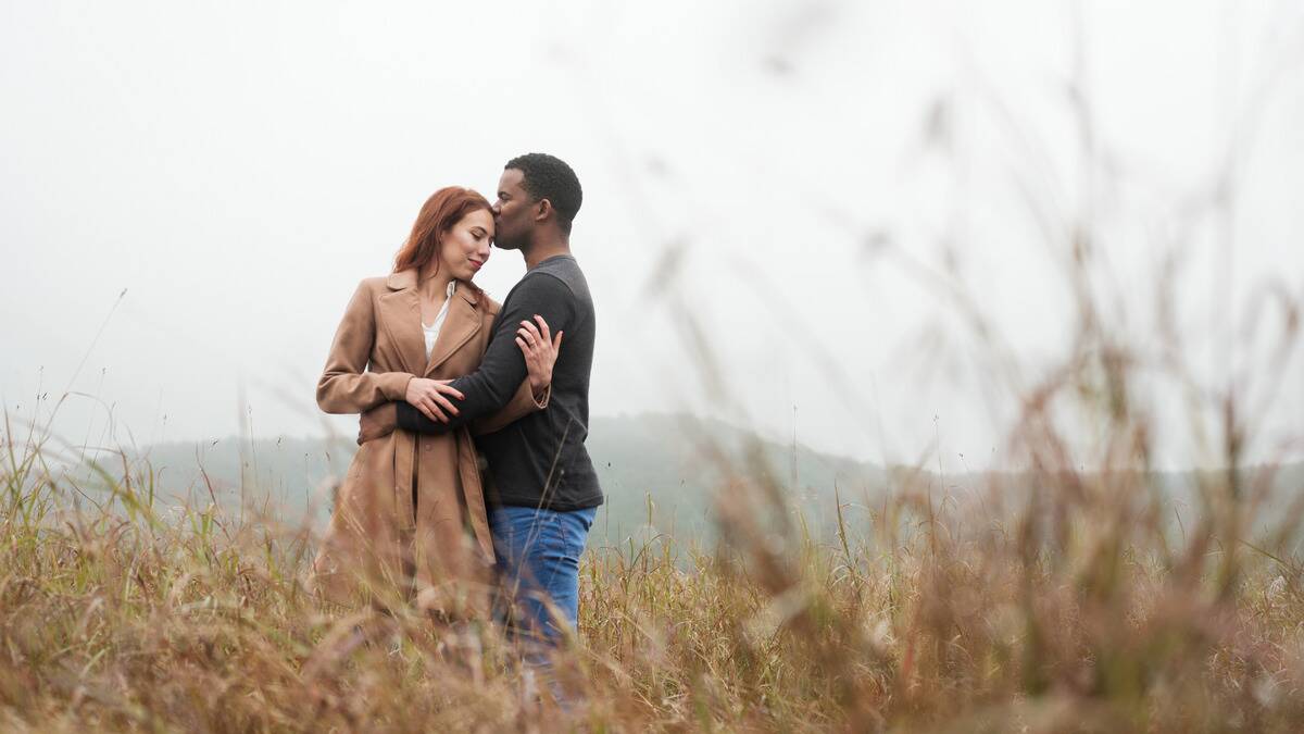 A couple standing in a field of tall grass, holding onto one another, the man pressing a kiss to the woman's forehead.