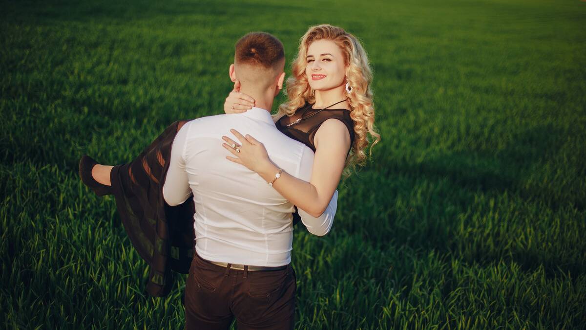 A man, faced away from the camera, carrying his girlfriend bridal style across a field as she smiles into the camera.