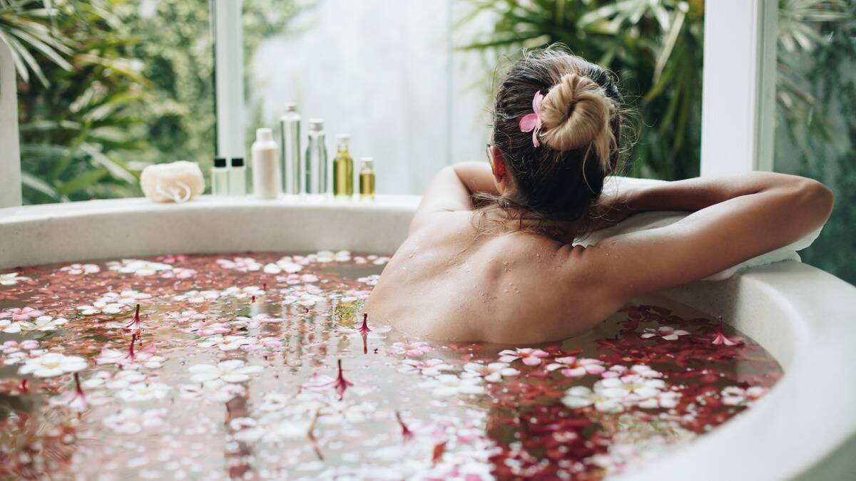 A woman in a bathtub full of flowers next to a window with some greenery outside. She's leaning her arms on the tub's edge, facing away from the camera.