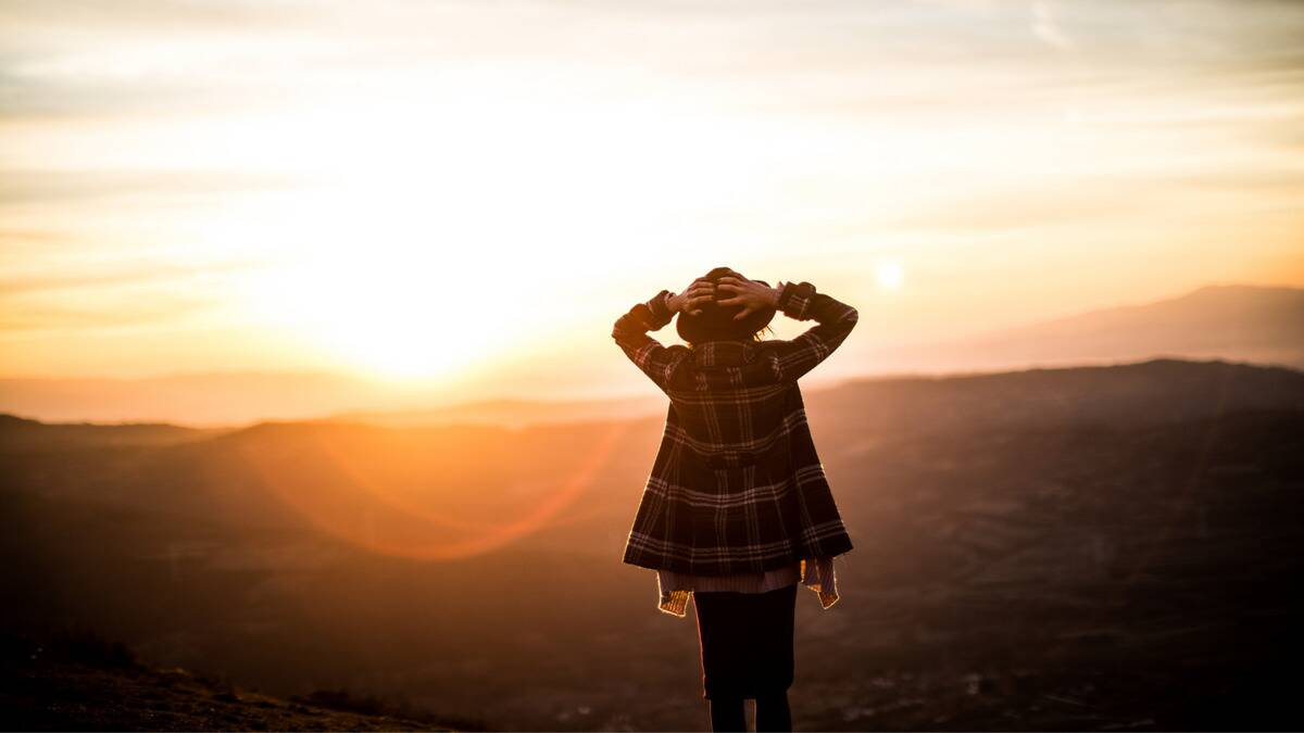 A woman standing against a horizon, hands on her head, looking out into the distance, faced away from the camera, the sun rising beside her.