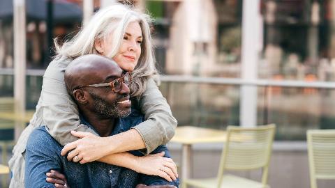 A woman standing behind her partner who's sitting down, wrapping her arms around his shoulders and resting her chin on his head as he turns to try and look at her.