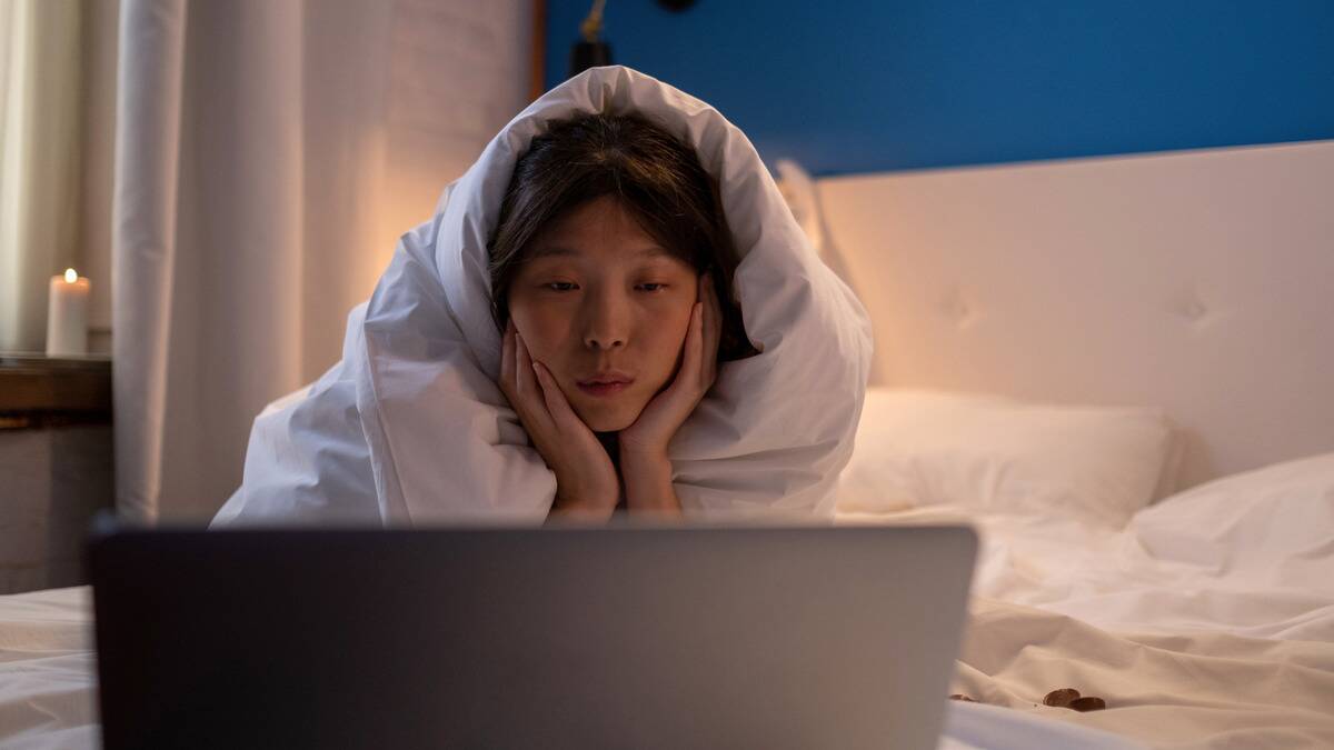 A woman laying on her stomach in bed, wrapped in a blanket, her chin in her hands, watching something on her laptop.