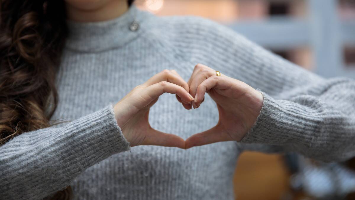 A torso shot of a woman in a grey sweater holding her hands in a heart shape over where her heart is in her chest.